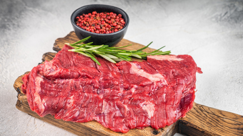 A raw inside skirt steak on a cutting board with pink peppercorns and rosemary