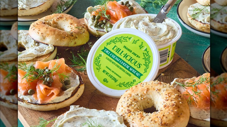 A tub of Dill•icious Cream Cheese Spread next to bagels and lox on a wooden cutting board with plates surrounding it.