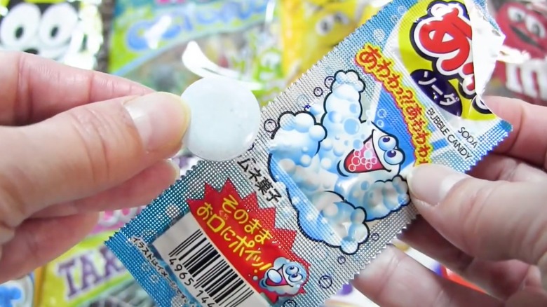 Hand holds an effervescent hard candy tablet in front of Coris Awa Soda Bubble Candy packaging