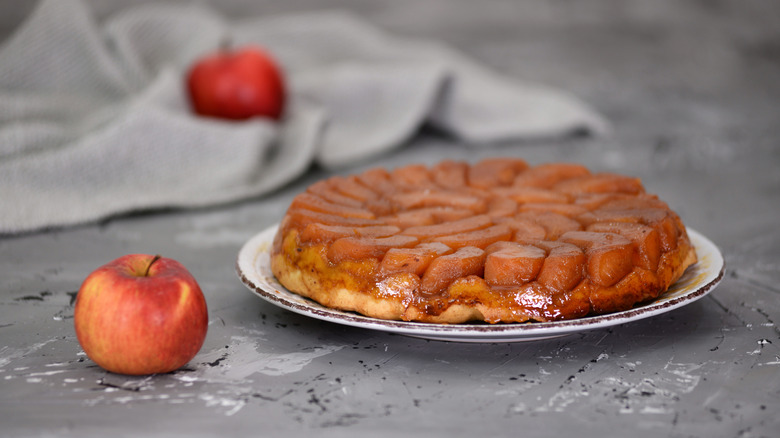 A whole tarte tatin against a grey background with apples next to the plate