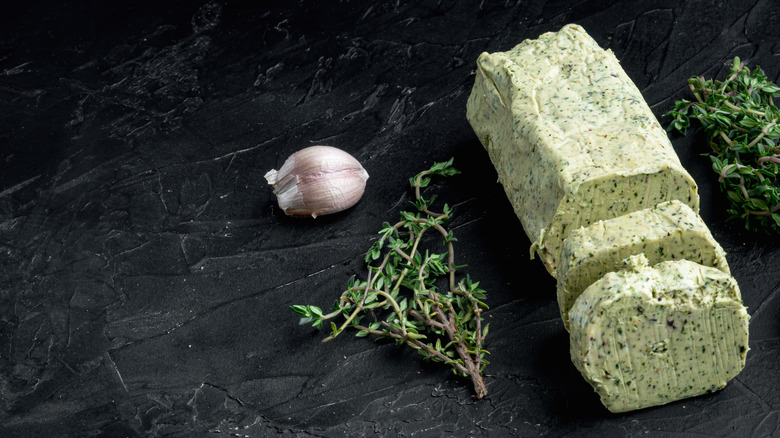 Stick of herb compound butter, with cloves of garlic and sprigs of herbs next to it