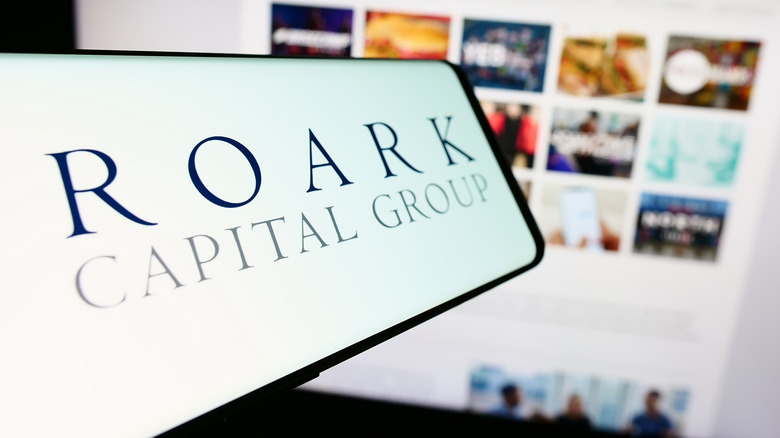 A phone reading "Roark Capital Group" in front of a blurry computer screen