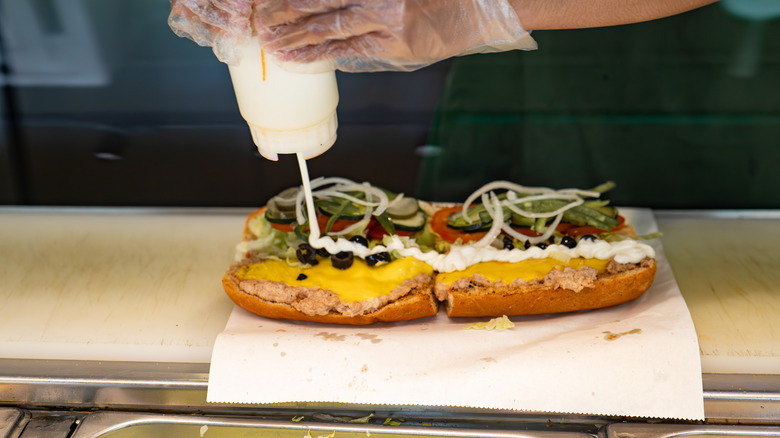 A Subway sandwich being prepared, with mayonnaise being poured onto a sandwich with tuna, cheese, olives, and salad