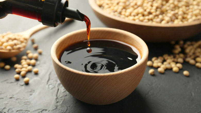Soy sauce is being poured from bottle into a bowl with soybeans in the background.
