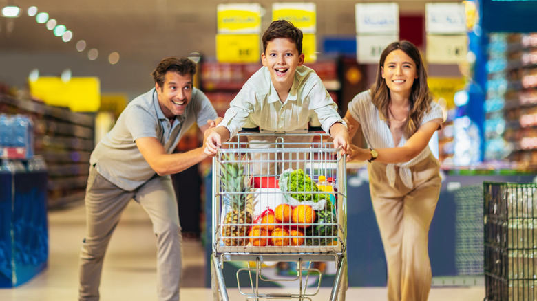Happy child kneeling on shopping card, with smiling mother and father pushing him down an aisle