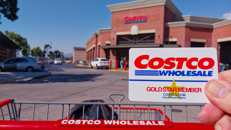 Person holding Costco Gold Star Member card up, in front of their shopping cart, with a Costco store in the background