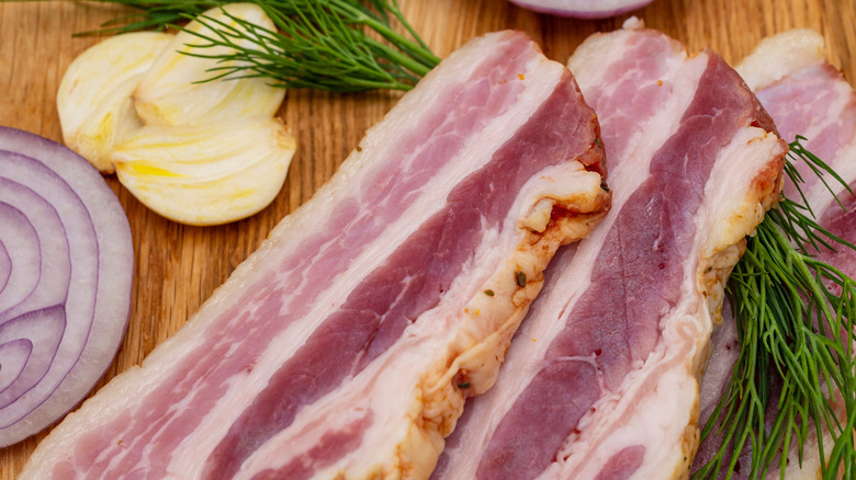 Slices of bacon on a wooden board, with sliced onions and herbs
