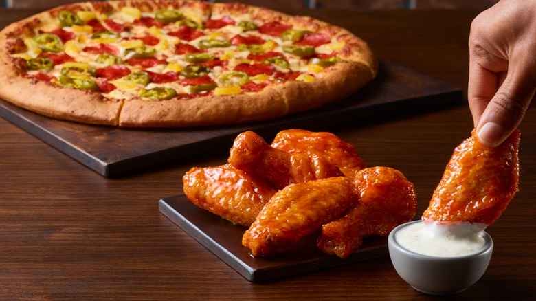 Front focus of Pizza Hut wings, one being dipped in sauce, with a blurred pizza in the background