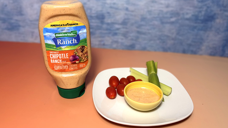 A bottle of chipotle ranch next to a plate with celery, tomatoes, and a bowl of dressing