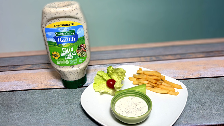 A bottle of green goddess ranch next to a plate with salad, fries, and a bowl of dressing