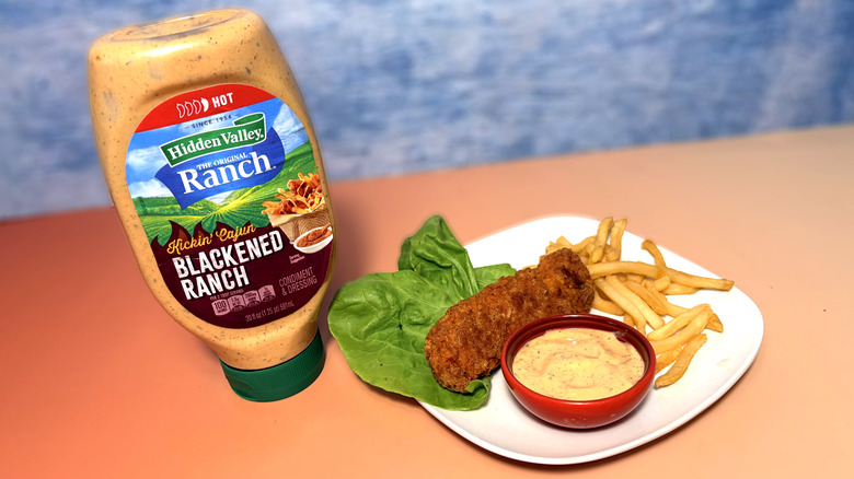 A bottle of blackened ranch next to a plate with salad, chicken tender, fries, and a bowl of dressing