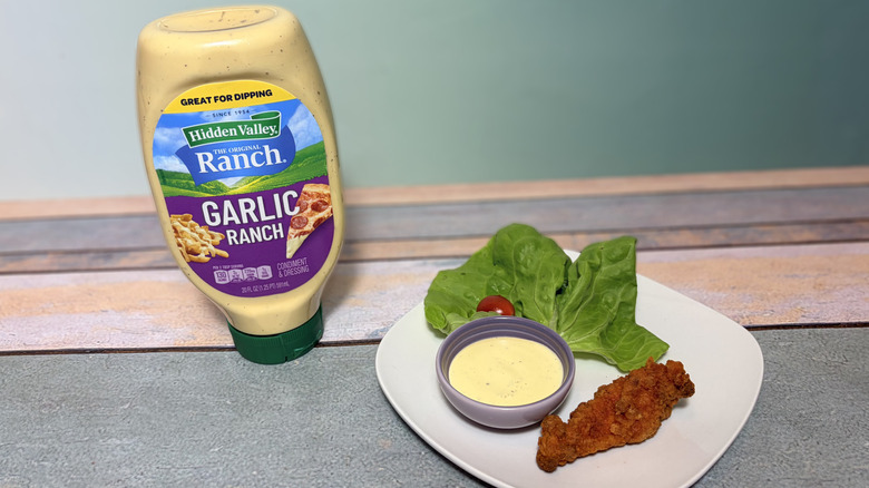 A bottle of garlic ranch next to a plate with salad, chicken tender, and a bowl of dressing