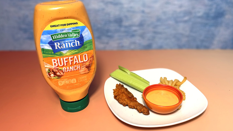 A bottle of buffalo ranch next to a plate with celery, chicken tender, fries, and a bowl of dressing