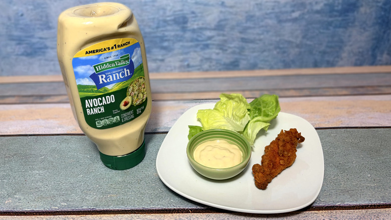 A bottle of avocado ranch next to a plate with lettuce, chicken tender, and a bowl of dressing