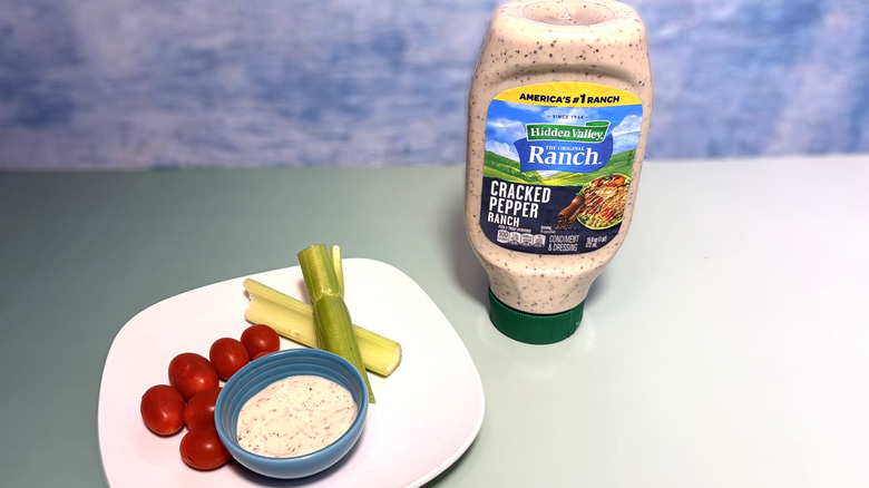 A bottle of cracked pepper ranch next to a plate with celery, tomatoes, and a bowl of dressing