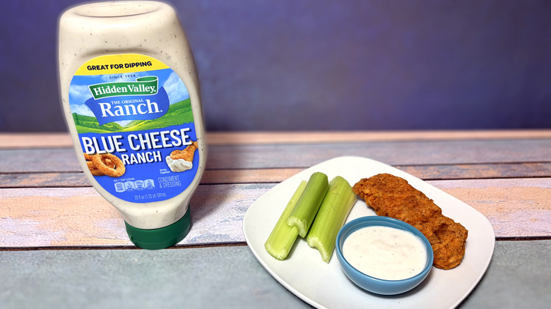 A bottle of blue cheese ranch next to a plate with celery, chicken tender, and a bowl of dressing