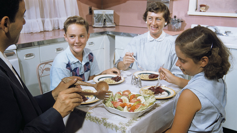 A vintage image of a family eating a meal in a small kitchen with pink walls, circa 1960