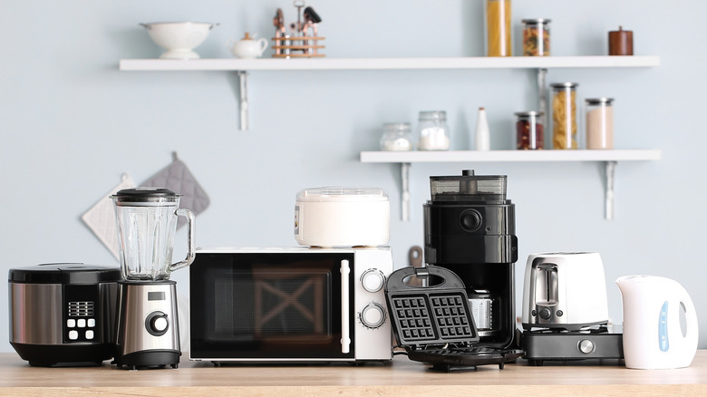 Various kitchen appliances on a wood countertop with shelves of items blurred in the background