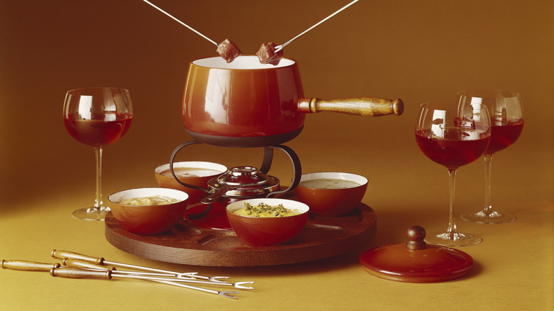 A vintage fondue set with sides of different cheeses and three glasses of wine