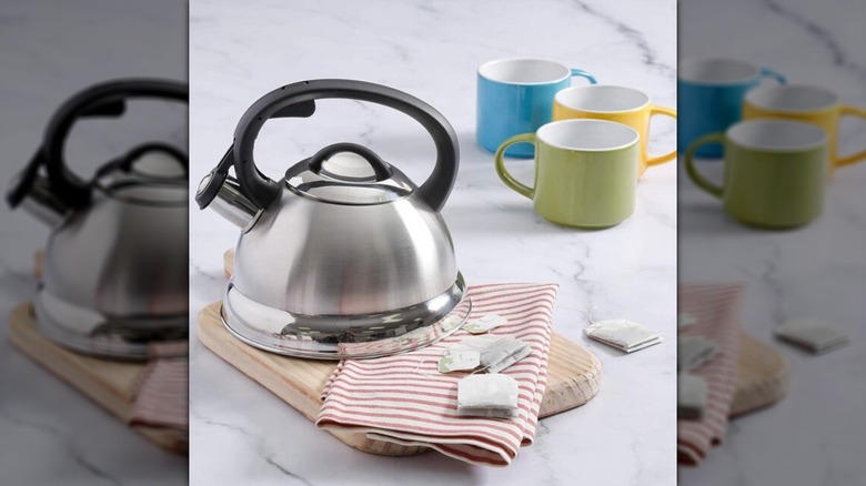 Mr. Coffee Flintshire Stainless Steel Kettle on top of a cutting board on a countertop next to three colorful mugs