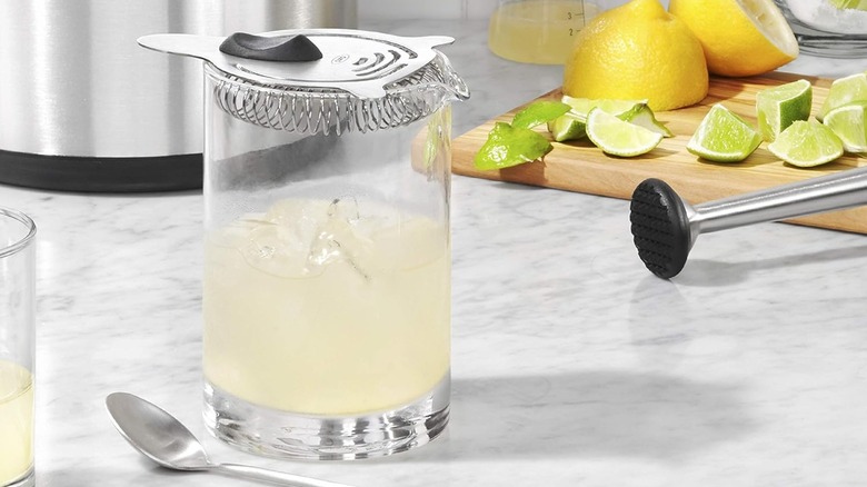 Close-up of the OXO Hawthorne strainer on a spouted glass filled with ice and lemon/lime juice with lemons, limes, and other cocktail tools on a counter