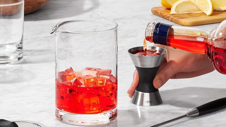 Pouring a mixer into the OXO jigger on the counter next to a spouted glass and other cocktail items