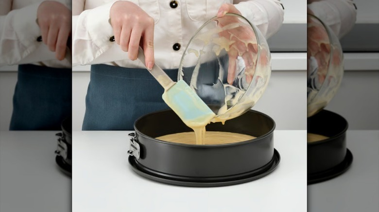 A person's hands uses a light blue spatula to pour cake batter from a glass bowl into a round pan.
