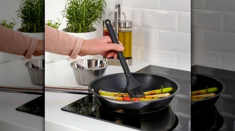 Hand holding silicone brush over vegetables in a pan on a glass cooktop in a kitchen.