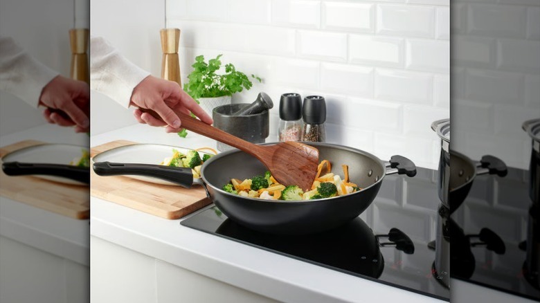 Hand holding a dark wood wok turner with wok full of veggies in kitchen with white subway tile.