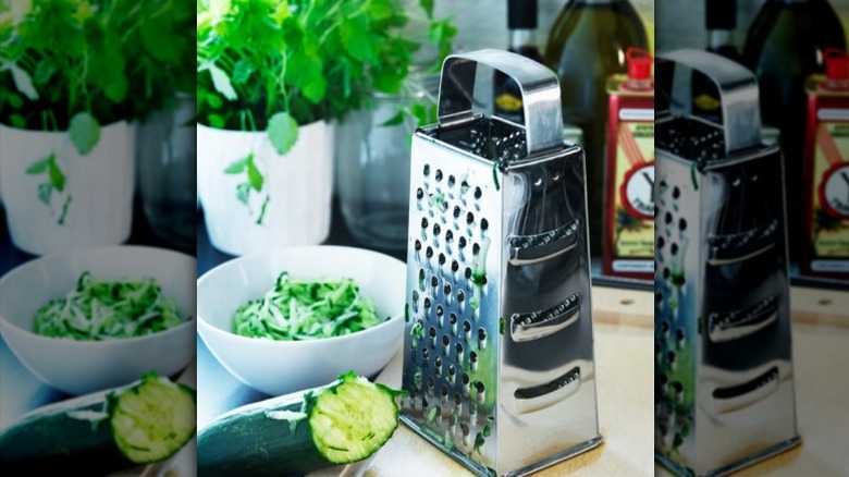 Stainless steel grater with zucchini on kitchen counter with white bowl full of grated zucchini behind it.