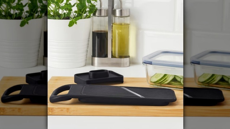 Black mandoline on light wood cutting board with a container of sliced cucumbers also on the counter.