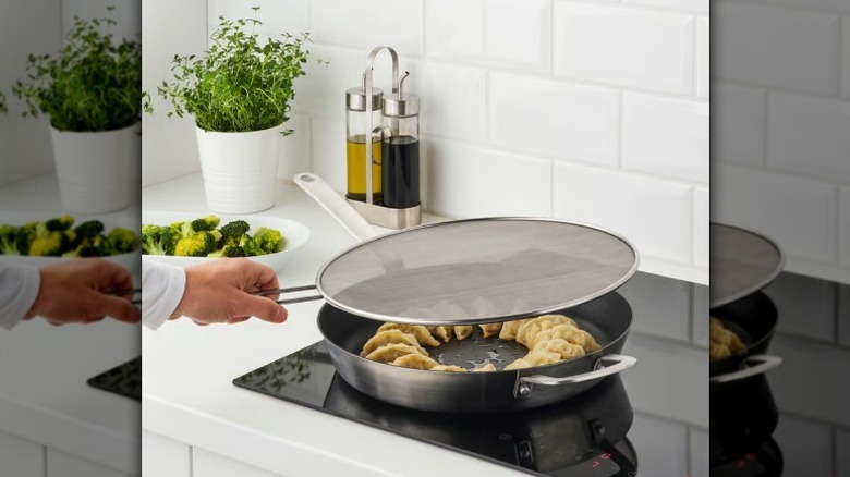 Hand holding splatter screen over pan with dumplings in kitchen with white subway tile backsplash.