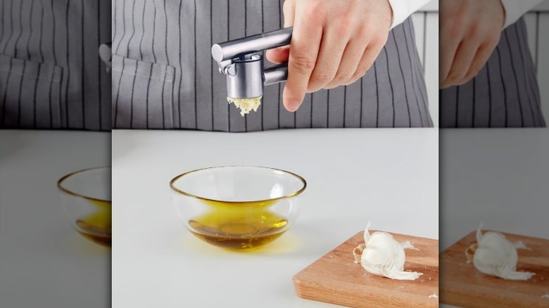Hand holding garlic press over a small glass bowl of oil with wooden cutting board with garlic on it nearby.
