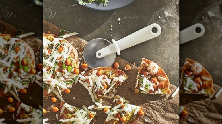 Pizza cutter with white handle rests on board with sliced pizza with cheese and chickpeas on top.