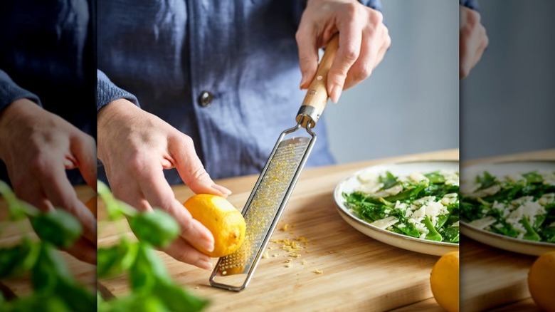 A person's hands zests a bright yellow lemon on a metal zester with wooden handle as a plate with salad on it sits beside.