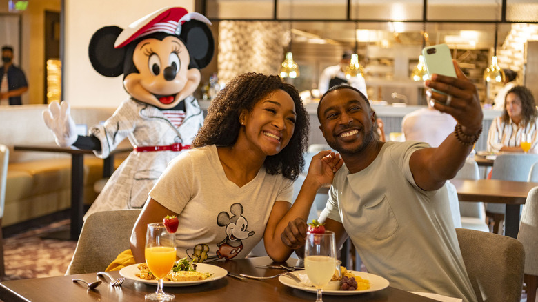 A couple takes a selfie in a Disney World restaurant with a costumed Minnie Mouse in the background
