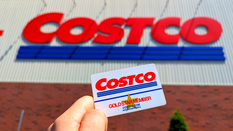 A hand holding a Costco card in front of a Costco storefront