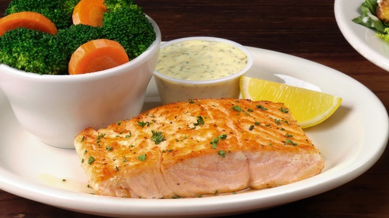 Texas Roadhouse salmon on a plate with tartar sauce, a lemon wedge, and a bowl of carrots and broccoli