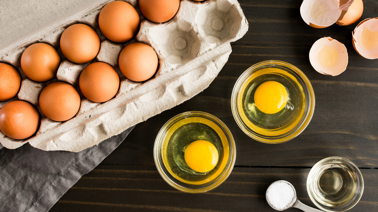 A carton of eggs on a table with two eggs cracked into small bowls