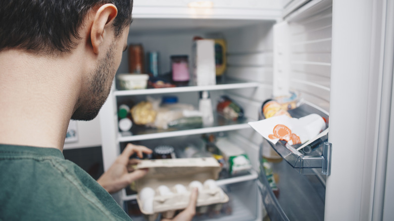 A man standing in the door of his refrigerator, inspecting a carton of eggs
