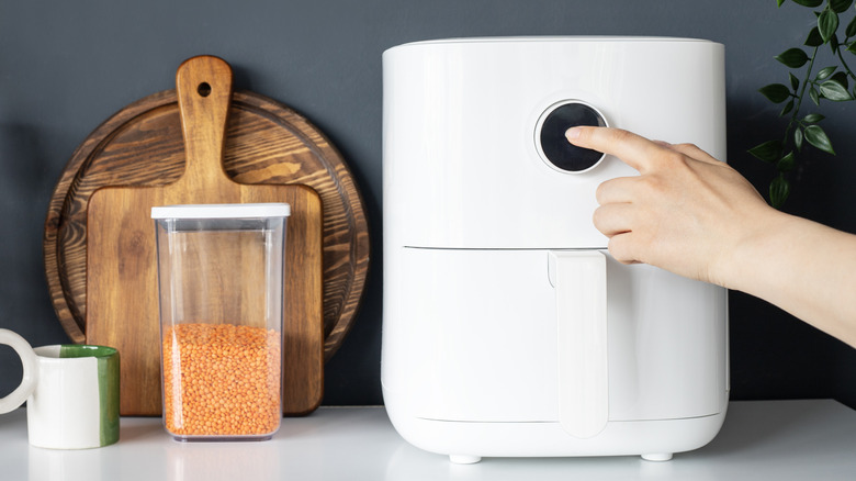A person using an air fryer placed on a kitchen countertop with chopping boards and a jar of raw lentils