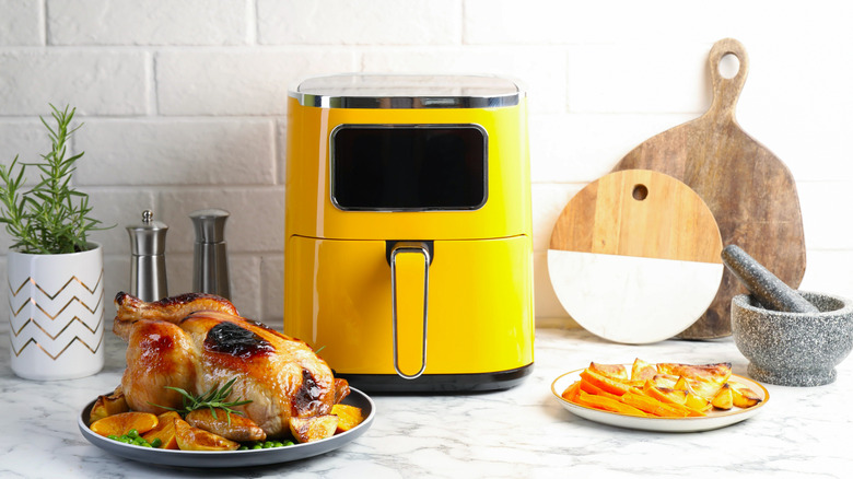 A yellow air fryer on a kitchen table with a whole roasted chicken and fries