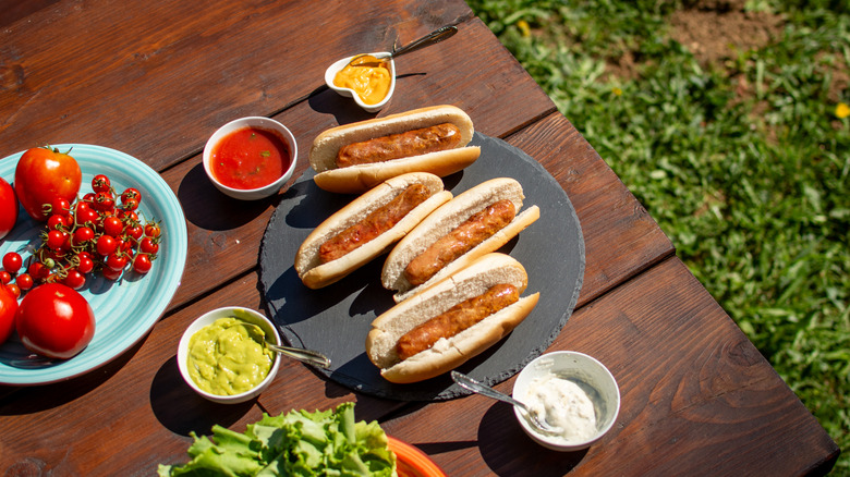 Four hot dogs on a picnic table outside, surrounded by various toppings
