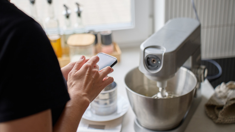A woman checks her phone while the stand mixer is mixing ingredients
