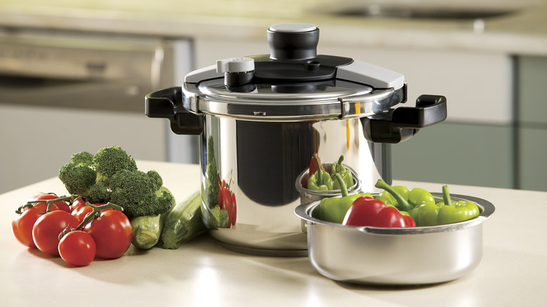 A silver and black pressure cooker on a counter next to fresh broccoli, tomatoes, and bell peppers