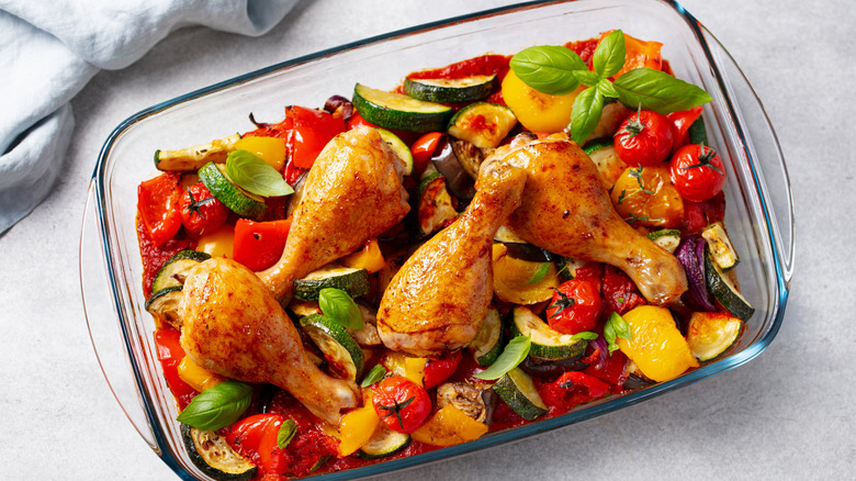 A glass dish filled with roasted chicken drumsticks and veggies on a countertop