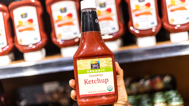 Hand holding up a 365 Organic tomato ketchup bottle at a Whole Foods store.