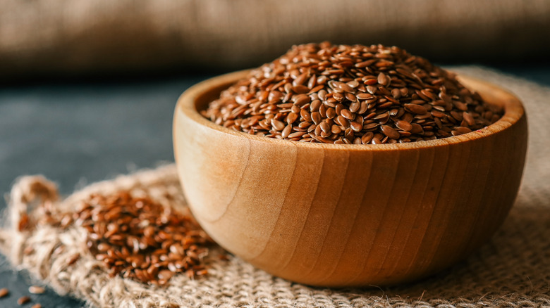 Focus on a wooden bowl of flaxseeds next to blurred flaxseeds on a rustic mat and background