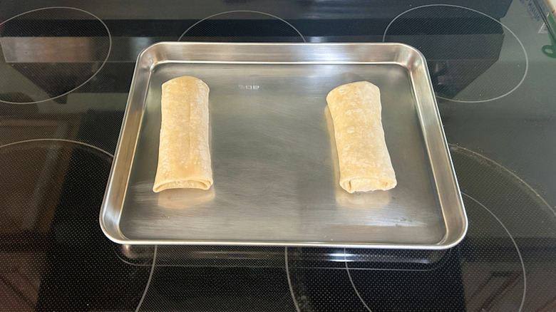 Two uncooked frozen chicken burritos in a metal pan on a stovetop