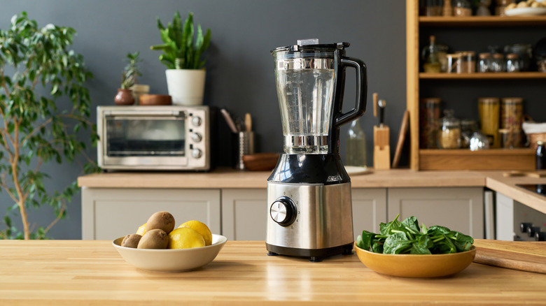 A blender on a wood countertop with kiwis, lemons, and spinach in bowls